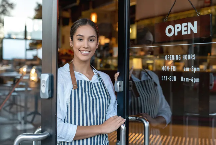 A happy cafe owner standing at the entrance of her shop under an open sign.